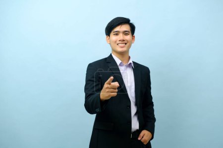 Foto de Portriat of asian young smart happy businessman dressed in suit standing straight, smiling, and pointing finger at camera with isolated light blue background. Business concept. - Imagen libre de derechos