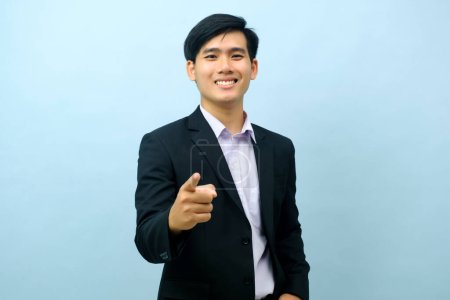 Foto de Portriat of asian young smart happy businessman dressed in suit standing straight, smiling, and pointing finger at camera with isolated light blue background. Business concept. - Imagen libre de derechos