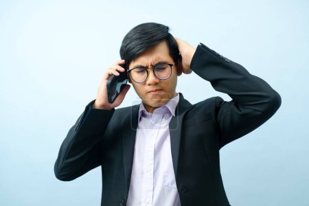 Foto de Portrait of young smart asian businessman in eyeglasses standing and talking on mobile phone stressed and frustrated about business with light blue isolated background. Business and connection concept. - Imagen libre de derechos