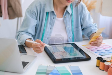 Foto de Young asian tailor designer designing on a new fashion collection by using tablet. She is sketching and drawing on a digital tablet at the table in her design studio. - Imagen libre de derechos