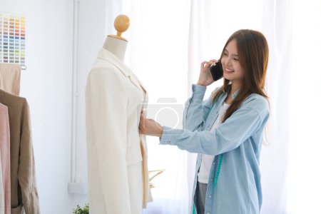 Foto de Young asian professional fashion designer is a working woman. She is standing and talking on mobile consulting client about her suit and about online order in fashion. She also give a consultation for - Imagen libre de derechos