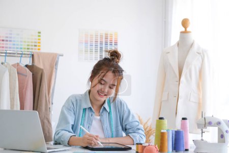Téléchargez les photos : Jeune tailleur asiatique dessinant sur une nouvelle collection de mode en utilisant une tablette. Elle dessine et dessine sur une tablette numérique à la table de son studio de design. - en image libre de droit