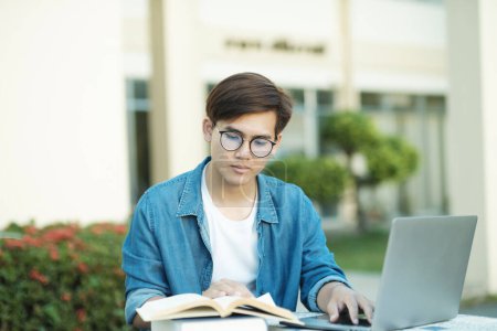 Photo for Young male college student with eyeglasses and in casual clothings sitting outdoor to study and read books using laptop for school project and research. Education and E-Learning concept. - Royalty Free Image