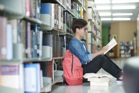 Photo for Young male college student wearing eyeglasses and casual clothings sitting on the floor reading book, studying and doing research for school project at a library. Learning and Educational concept. - Royalty Free Image