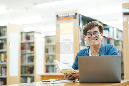 Photo for Young male college student wearing eyeglasses and in casual cloths sitting at desk looking at camera to study and read books using laptop at library for research or school project. Learning concept. - Royalty Free Image