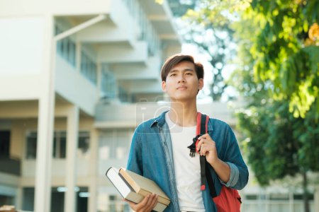 Photo for Young smart motivated male collage student in casual clothings and backpack standing outdoor with campus building in the backfround and holding books while looking up ready for study. Education concept. - Royalty Free Image