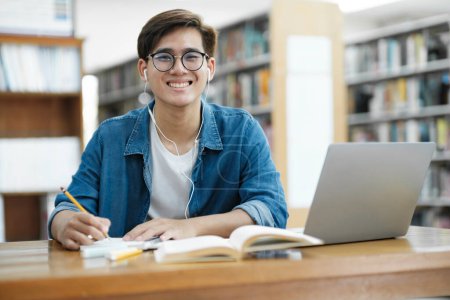 Photo for Young male college student wearing eyeglasses and in casual cloths sitting at desk studying, reading book, and writing down notes using laptop wearing headphones looking at camera at library for research or school project. E-Learning concept. - Royalty Free Image