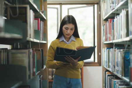 Photo for Young female college highschool student in casual outfit standing, reading or choosing books to read at library for studying, academic research, school work, project, or exam. Learning, Education, Library concept. - Royalty Free Image