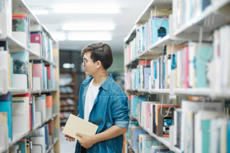Photo for Young male student in casual outfit with backpack standing, looking and choosing books to read at library for studying, academic research, or school work. Learning, Education concept. - Royalty Free Image