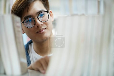 Photo for Close up of young male student in casual outfit with backpack standing, looking and choosing books to read at library for studying, academic research, or school work. Learning, Education concept. - Royalty Free Image