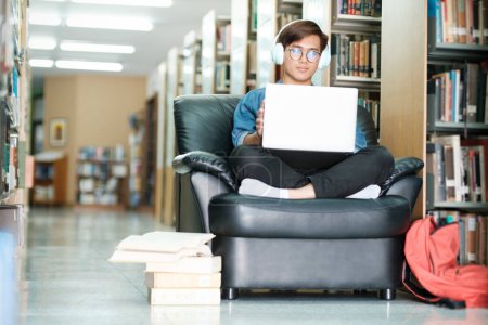 Photo for Young male college student wearing eyeglasses and in casual cloths sitting on couch, studying, and using laptop wearing headphones at library for research or school project. E-Learning and Education concept. - Royalty Free Image