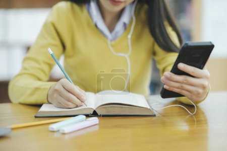 Photo for Young female college highschool student with headphones and in casual cloths sitting at desk studying, reading books, and writing with handphone at library for research or school project. E-Learning Education Library concept. - Royalty Free Image