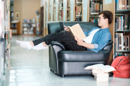 Foto de Joven estudiante universitario con anteojos y ropa casual sentado en el sofá leyendo un libro, estudiando e investigando para un proyecto escolar en una biblioteca. E-Learning y concepto educativo. - Imagen libre de derechos