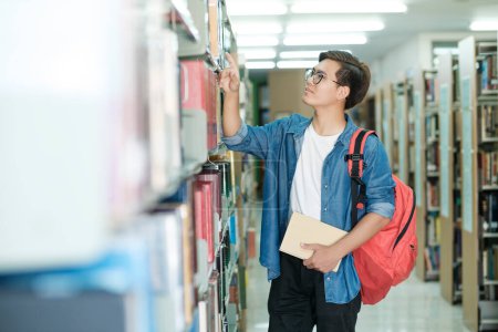 Photo for Young male student in casual outfit with backpack standing, looking and choosing books to read at library for studying, academic research, or school work. Learning, Education concept. - Royalty Free Image
