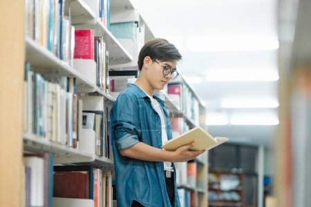 Photo for Young male student in casual outfit with backpack standing, reading or choosing books to read at library for studying, academic research, or school work. Learning, Education concept. - Royalty Free Image