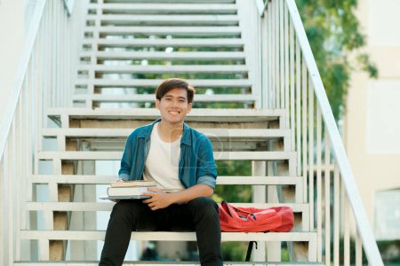 Photo for Young male college student in casual clothings looking at camera, holding books, and sitting outdoor on stairs to study and read books preparing for school project and research. Education concept. - Royalty Free Image