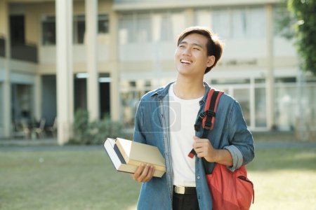 Photo for Young smart motivated male collage student in casual clothings and backpack standing outdoor with campus building in the backfround and holding books while looking up ready for study. Education - Royalty Free Image
