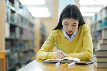 Photo for Young female college highschool student in casual cloths sitting at desk studying, reading books, and writing alone at library for research or school project. E-Learning Education Library concept. - Royalty Free Image