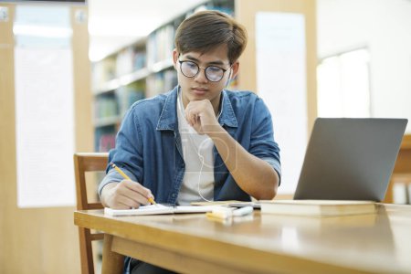 Photo for Young male college student wearing eyeglasses and in casual cloths sitting at desk studying, reading book, thinking hard, and writing down notes using laptop wearing headphones at library for research - Royalty Free Image