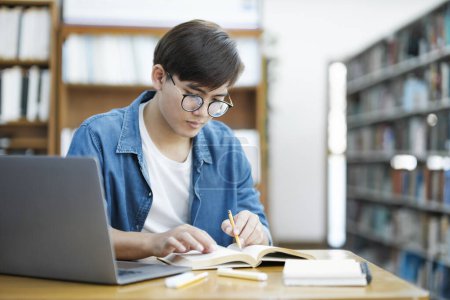 Photo for Young male university college student wearing eyeglasses and in casual cloths studying, reading, and writing down notes using laptop on desk at library for school project or exam preparation. E - Royalty Free Image
