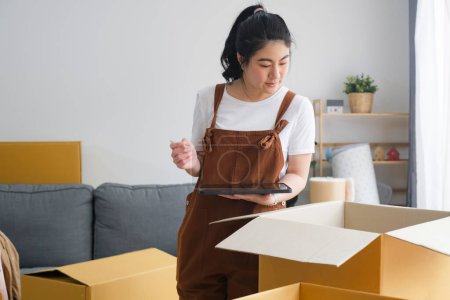 Foto de La mujer asiática que revisa y que embala la caja de cartón se prepara para moverse al nuevo envío de la reubicación de la casa o comprueba el paquete de mercancías antes de la entrega al cliente de compras en línea por lista de comprobación - Imagen libre de derechos