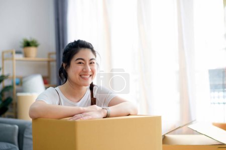 Foto de Mujer joven sonriente moviéndose en su nueva casa y apoyándose en una caja de cartón
. - Imagen libre de derechos