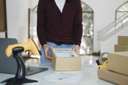 Photo for Close up of young female online business, store owner or entrepreneur standing at desk with laptop, scanner, and cardboard boxes holding and putting label tag on parcel for preparation before shipment - Royalty Free Image