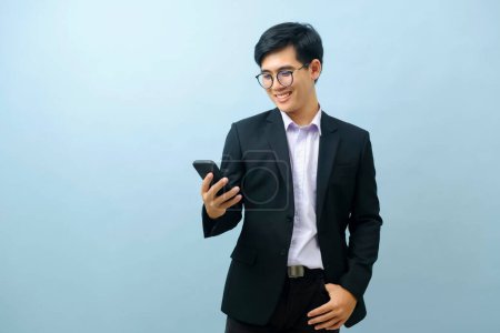 Photo for Portrait of young smart asian businessman standing and smiling while using smartphone with light blue isolated background. Business, connection concept. - Royalty Free Image