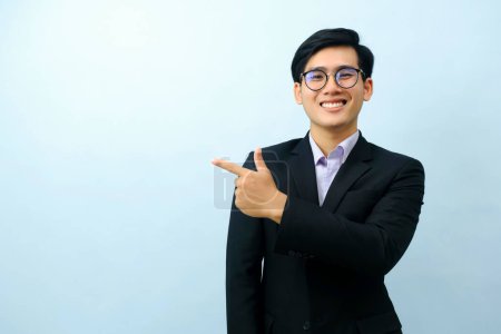 Téléchargez les photos : Portriat of asian young smart happy businessman dressed in suit standing straight, smiling, and pointing finger looking at camera with isolated light blue background. Business concept. - en image libre de droit