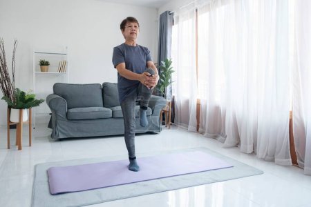Téléchargez les photos : Mode de vie sain et concept de vieillissement. Équilibre dans la vie. Femme âgée faisant des exercices d'étirement yoga dans le salon à la maison. - en image libre de droit