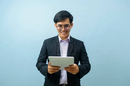Foto de Retrato de un joven empresario asiático inteligente con gafas de pie y sonriendo mientras usa la tableta para trabajar o comunicarse con un fondo aislado de color azul claro. Negocios, concepto de conexión. - Imagen libre de derechos
