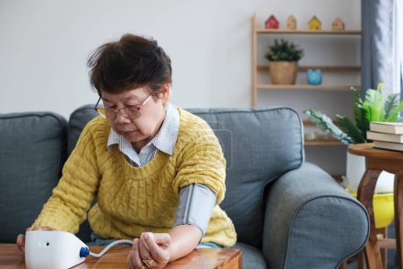 Foto de Problema de hipertensión en la tercera edad. Mujer asiática midiendo la presión arterial arterial usando el manguito del esfigmomanómetro sentado en el sofá en casa. Asistencia sanitaria, problemas de salud. - Imagen libre de derechos