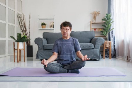 Foto de Una mujer mayor encuentra paz interior a través del yoga y la meditación en casa, abrazando la relajación y la tranquilidad de la mente. Calma descalza hembra de mediana edad sentada en la alfombra en la postura de loto medio, haciendo - Imagen libre de derechos