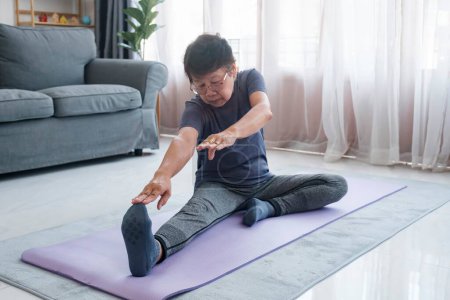 Foto de Mujer madura activa haciendo ejercicio de estiramiento en la sala de estar en casa. Ajuste señora estirando los brazos y la espalda mientras está sentado en la esterilla de yoga. - Imagen libre de derechos