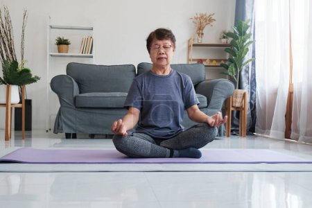 Téléchargez les photos : Une femme âgée trouve la paix intérieure par le yoga et la méditation à la maison, embrassant la relaxation et la tranquillité de l'esprit. Calme pieds nus femme d'âge moyen assis sur le tapis en demi-lotus posture, ce qui rend - en image libre de droit