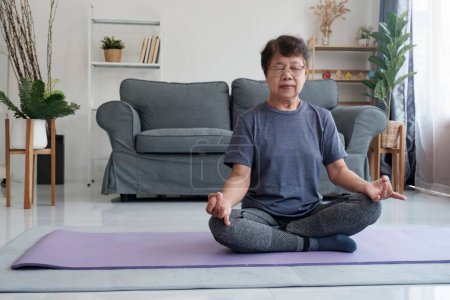 Foto de Una mujer mayor encuentra paz interior a través del yoga y la meditación en casa, abrazando la relajación y la tranquilidad de la mente. Calma descalza hembra de mediana edad sentada en la alfombra en la postura de loto medio, haciendo - Imagen libre de derechos