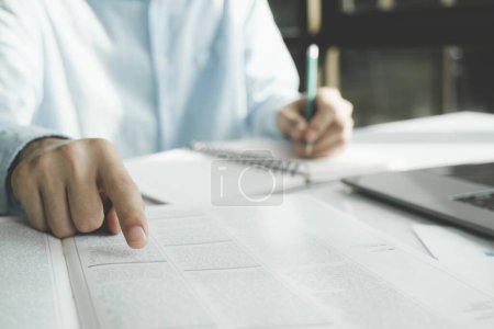 Photo for Closeup student hand writing essay in copy book at workplace table, making notes, summary from open book, doing school, college home task, research study - Royalty Free Image