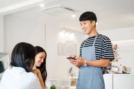 Photo for Cheerful smiling waiter in apron using table while take order and talk to clients cafe coffee shop visitors, friendly professional man server wear apron write menu choice, serving staff good customer - Royalty Free Image