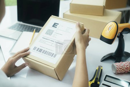 Photo for Close up of young female online business owner sitting at desk with laptop and boxes and putting shipping label on cardboard boxes or parcel of customers order preparing for shipment and delivery. - Royalty Free Image