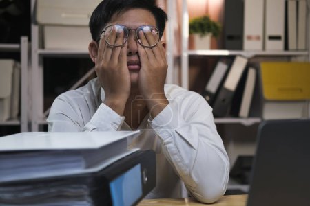 Photo for Exhausted young man with laptop in office working late sitting on desk in office overtime at night. Frustrated young businessman massaging his nose and keeping eyes closed while sitting at his working - Royalty Free Image
