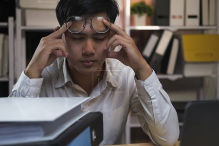 Photo for Exhausted young man with laptop in office working late sitting on desk in office overtime at night. Frustrated young businessman massaging his nose and keeping eyes closed while sitting at his working - Royalty Free Image