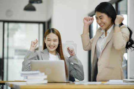 Téléchargez les photos : Deux femmes d'affaires extatiques célébrant un moment réussi au travail avec un ordinateur portable et des documents sur le bureau.. - en image libre de droit
