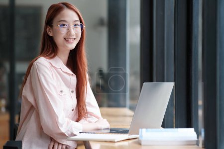 Téléchargez les photos : Charmant et confiant jeune étudiant universitaire ou femme d'affaires assis sur ordinateur portable dans le bureau - en image libre de droit