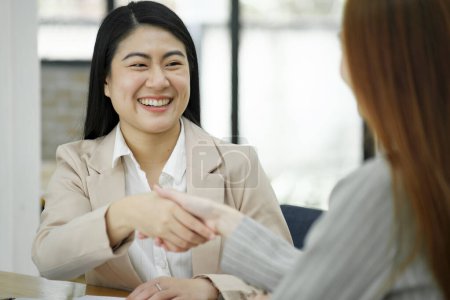 Foto de Dos mujeres de negocios sonrientes dándole la mano a un escritorio en la oficina, simbolizando un acuerdo exitoso o asociación. - Imagen libre de derechos