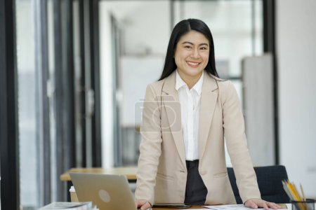 Foto de Una empresaria profesional y confiada sonriendo a la cámara, de pie junto a su escritorio con un portátil en una oficina moderna. - Imagen libre de derechos