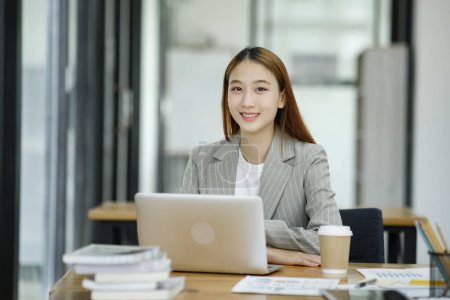 Eine professionelle und selbstbewusste Geschäftsfrau lächelt in die Kamera und steht mit einem Laptop an ihrem Schreibtisch in einem modernen Büro.