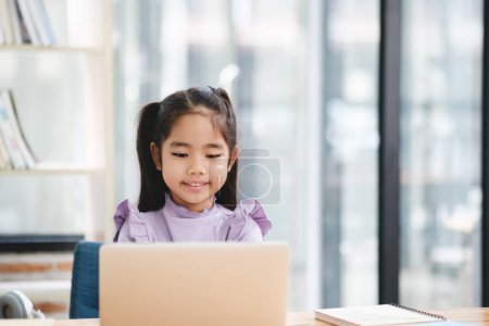 Photo for A young girl is sitting at a desk with a laptop in front of her. She is smiling and she is enjoying her time. The room is filled with books - Royalty Free Image