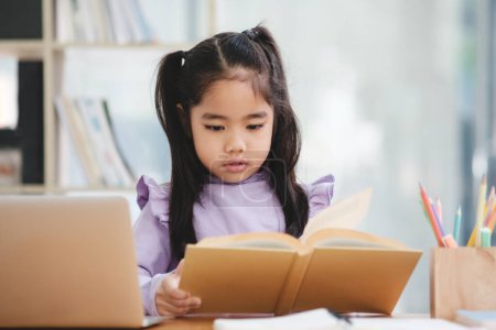 Photo for A young girl is sitting at a desk reading a book. She is wearing a purple shirt and has her hair in pigtails. The scene is set in a library or a classroom, with a laptop and a pencil on the desk - Royalty Free Image