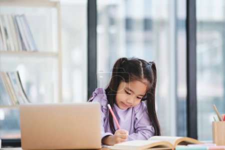 Photo for A young girl is sitting at a desk with a laptop and a book. She is writing on the laptop and she is focused on her work - Royalty Free Image