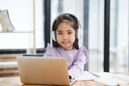Téléchargez les photos : Une jeune fille est assise à un bureau avec un ordinateur portable et un cahier. Elle porte des écouteurs et sourit. La scène suggère qu'elle est engagée dans une certaine forme d'apprentissage ou d'étude - en image libre de droit